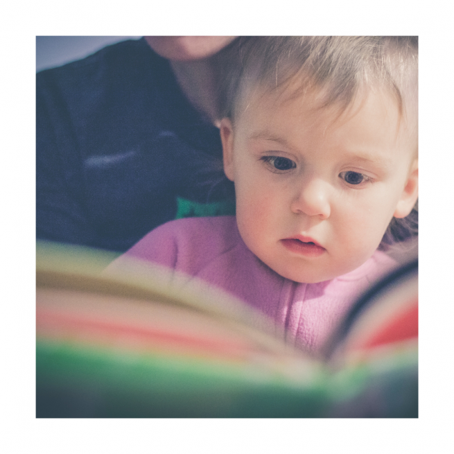 Kleinkind mit Buch und Bezugsperson im Hintergrund